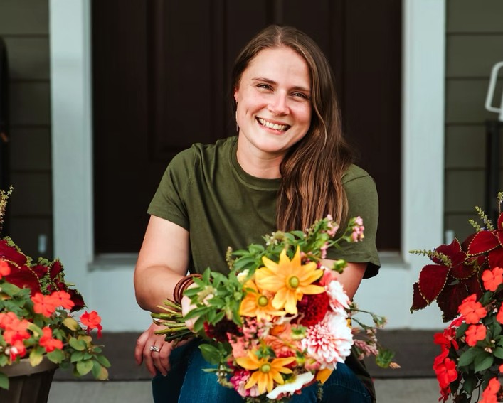 A portrait photo of a person sitting on a porch, smiling at the camera while holding a large bouquet of colorful flowers. There are large bronze planters planted with various colorful annual flowers.