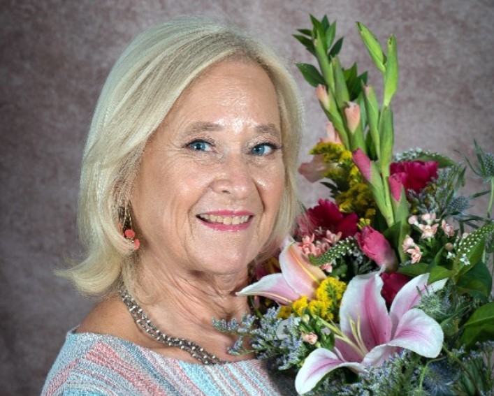 A person with bright blond hair is smiling at the camera while holding a large bouquet of colorful flowers next to their face.