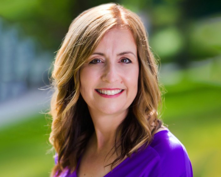 A portrait photo of a person with long auburn hair smiling at the camera on a sunny lawn.