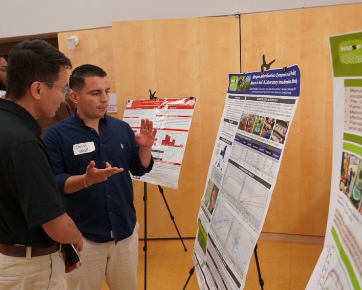 Two men talk near a research poster. The man on the right gestures with his hands.