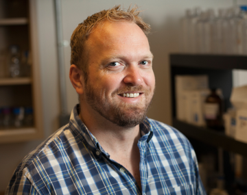 a man with red hair and a red beard and a checkered shirt stands in a lab