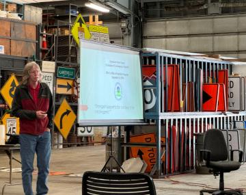 Person doing a presentation in a warehouse next to racks of traffic signs.