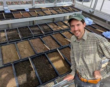person posing in front of a greenhouse full of trays of soils of different shades