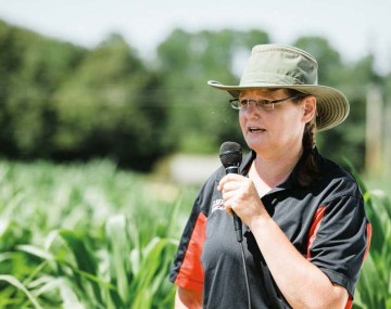 Person in a wide brimmed hat holding a microphone and standing in a green field of plants
