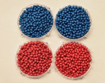 Four clear petri dishes arranged in a grid, displaying examples of insecticide seed treatments. The top two dishes are filled with pelletized seeds that have a bright blue coating, and the bottom two dishes contain pelletized seeds with a bright red coating.