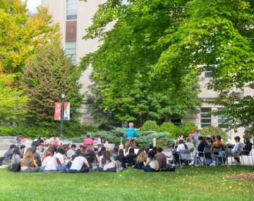 a crowd of students sit in front of building listening to an old man talk