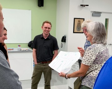 A group of 6 smiling people stand in a circle while one person holds their greenhouse map or diagram in the center of the circle, showing it to the group.