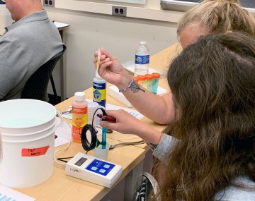 Two people hold electronic instruments in small cups of fertilizer mixture to measure PH and total dissolved solids, respectively.