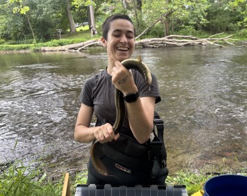 Photograph of a person with a buzz cut smiling holding a large brown and green American eel at the edge of a river.