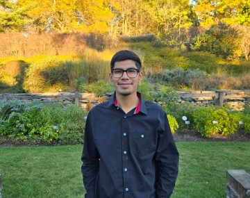 person with short dark hair and glasses stands in front of grass and trees with colorful leaves