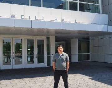 person with short black hair and gray shirt stands with hands in pockets