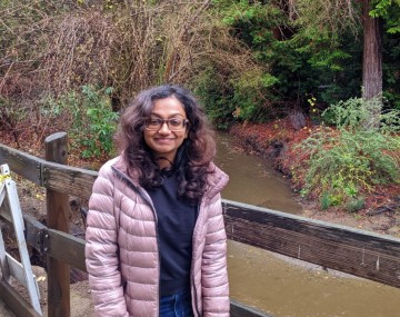 person with brown shoulder-length hair and lavender coat standing on bridge