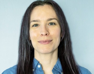 headshot of person with long dark hair in front of light blue background wearing blue button down shirt