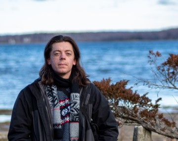 person with shoulder-length brown hair stands in front of body of water wearing dark coat and blue and red scarf
