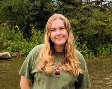 Student with long blond hair standing by the river, smiling