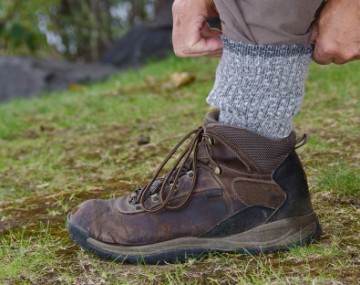 A close-up of a person outdoors adjusting their gray wool sock above a brown hiking boot on grassy ground.