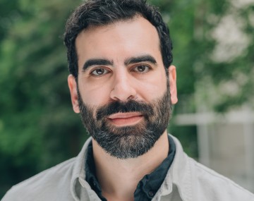 Bearded man wearing a light button-down shirt, standing outdoors with trees softly blurred in the background.