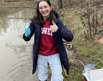 Photograph of a young woman standing in a stream holding a test tube.