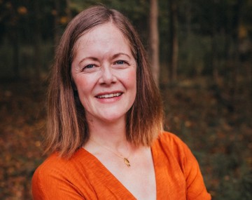Professional portrait of person standing with crossed arms and long brown hair wearing an orange shirt.
