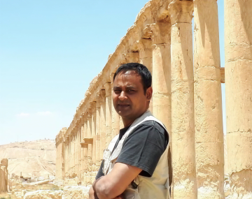 Man standing with arms crossed beside a row of ancient stone columns in a desert setting.
