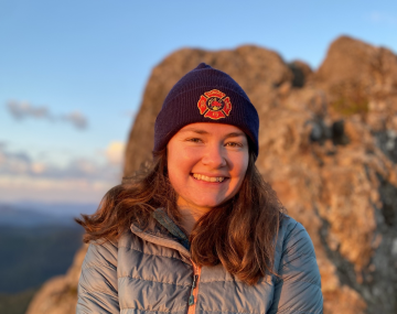 Photograph of a young woman in a jacket and wearing a beanie with a rocky shoreline behind her.