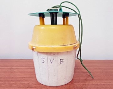 A squash vine borer bucket trap used to capture and monitor adult moth populations. The whole trap is slightly smaller than a 5 gallon (18.93 liters) bucket. The trap consists of a white base, a yellow top and an elevated green lid above the yellow top. There is a hole in the center of the yellow top that allows for SVB moths to enter the trap and be captured. The trap has a string attached to the elevated lid so it can be hung above the ground. 