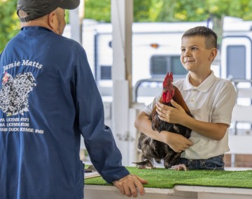 a boy holds a rooster on a table facing a man with his back to the camera