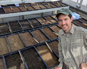 A smiling man wearing a green cap and a plaid button-down shirt stands in a brightly lit greenhouse. He rests one hand on the edge of a metal grating table that holds dozens of black rectangular planting trays. The trays are filled with varying shades of brown soil, and several contain small wooden marker stakes and newly sprouted tiny green seedlings.