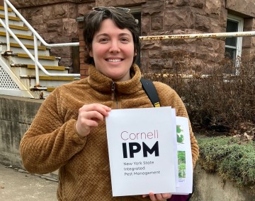 A smiling woman with short dark hair stands outdoors in front of a stone and brick building, holding up a booklet titled 'Cornell IPM: New York State Integrated Pest Management.' She wears a brown fleece jacket, light-colored pants, and sunglasses pushed up on her head. Behind her to the left are concrete steps with a white metal railing, and to the right, a low wall with bare shrubs.