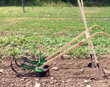 A manual seeder or push planter viewed from the side, parked in freshly agitated light brown soil.