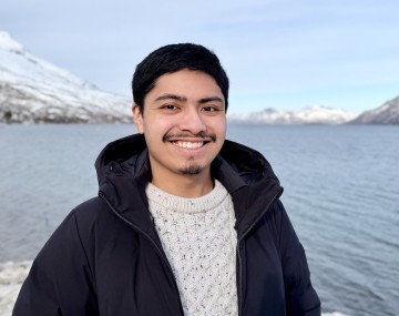 short dark hair, facial hair, white sweater, black jacket in front of lake and mountains