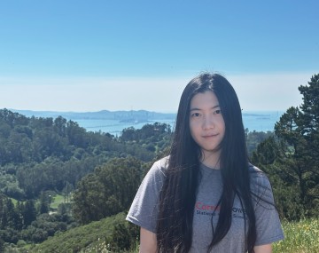 headshot of person with long dark hair wearing gray t-shirt in front of blue sky and mountains with trees on them