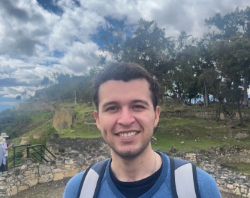 person with short dark hair, blue shirt, gray backpack straps by a ruins with pretty clouds