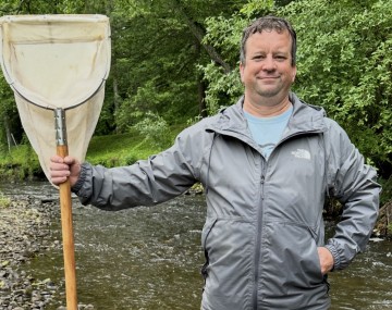 Photograph of a man in a rain jacket holding a large net standing in a stream.