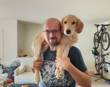 bald person with glasses and black t shirt with white writing holding a golden retriever puppy over his shoulders